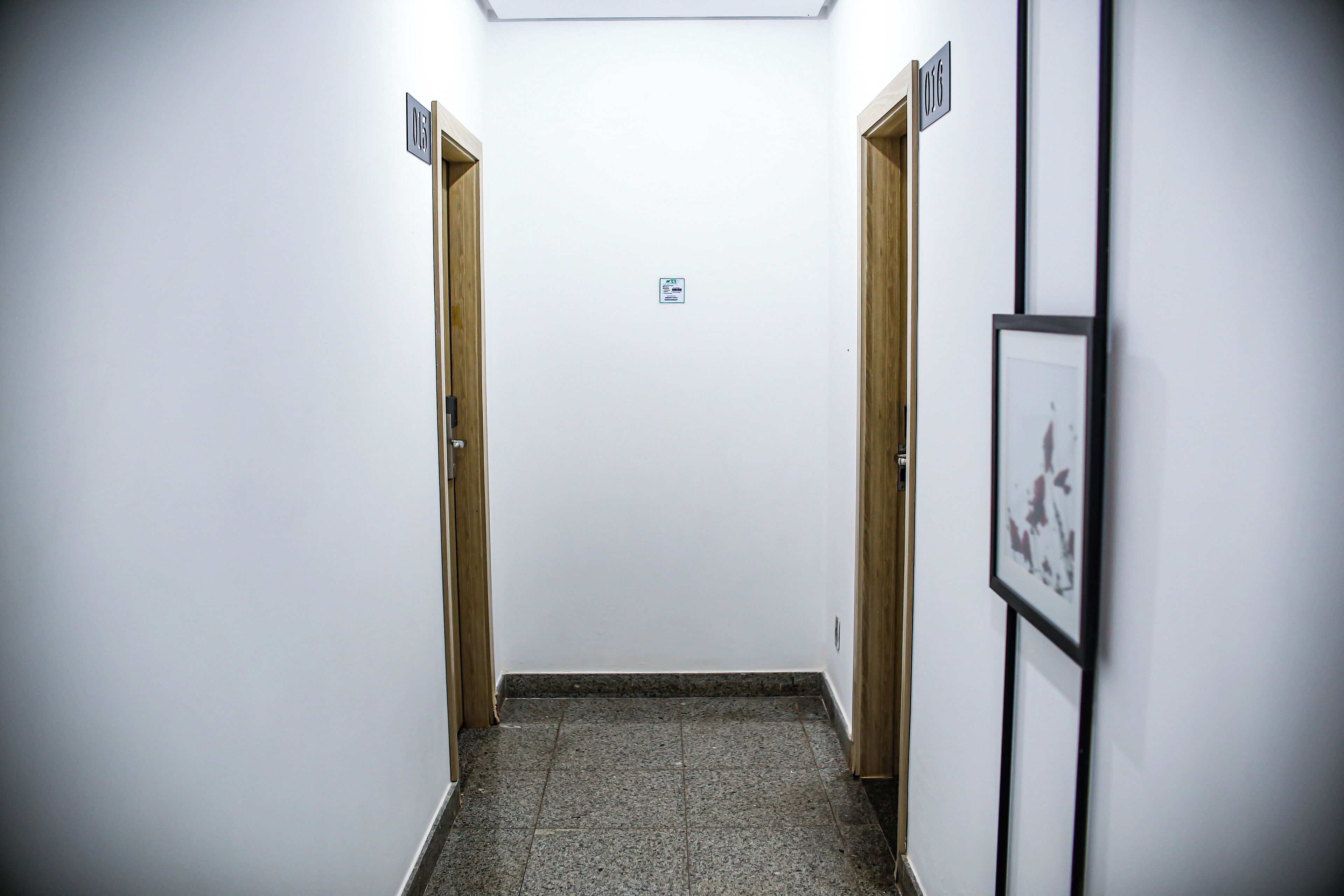 A narrow hallway with wooden doors on either side, a tiled floor, and a framed picture on the right wall.