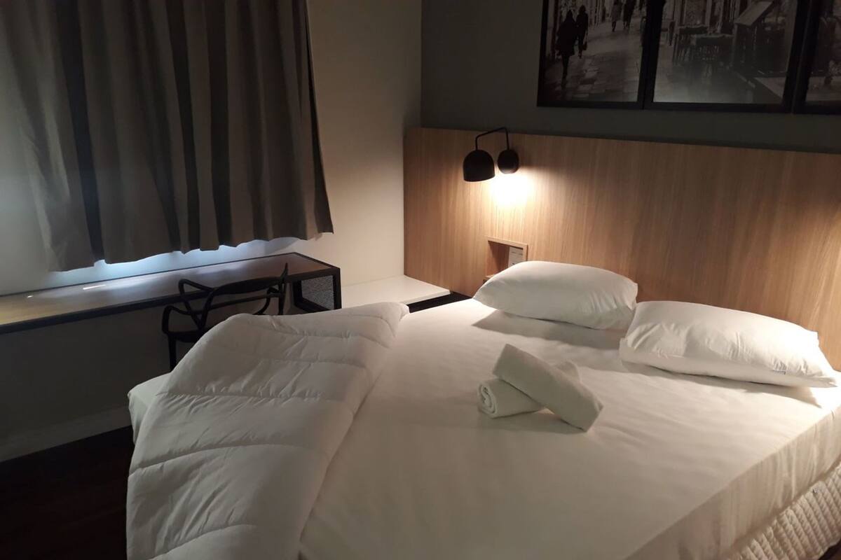 Un lit bien fait, avec du linge de lit blanc et des oreillers, un petit bureau avec une chaise, une lampe fixée au mur et un tableau encadré au-dessus du lit.