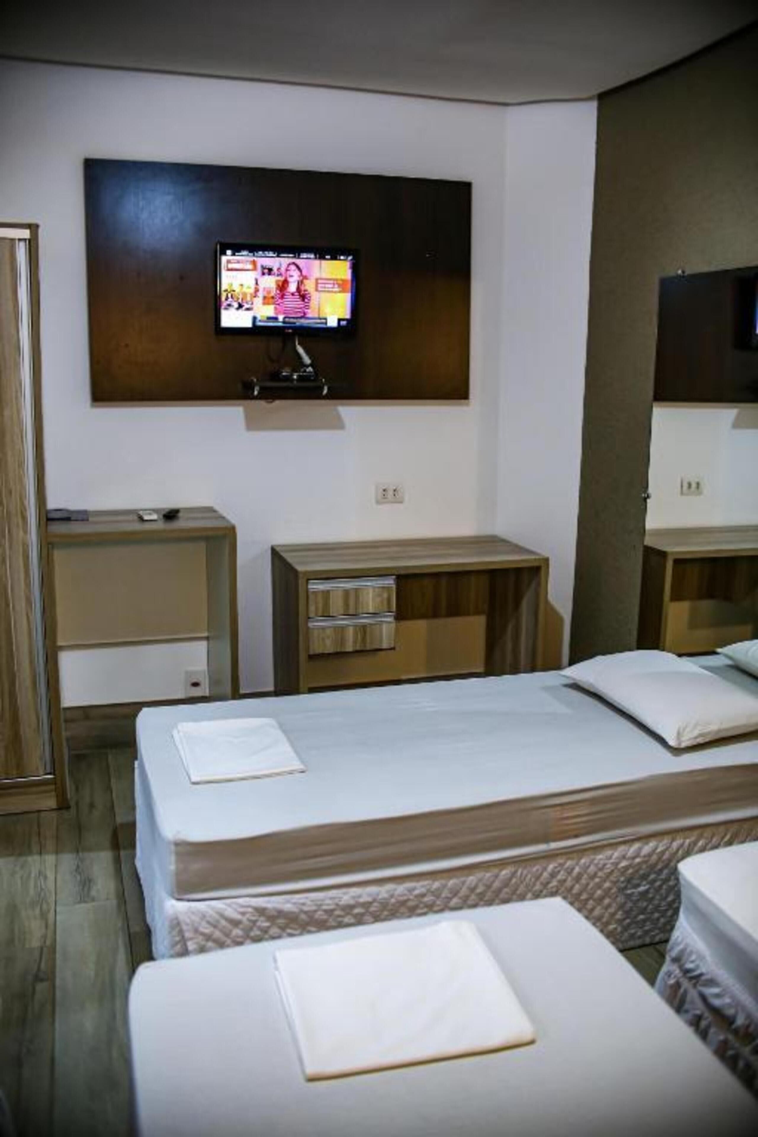 A hotel room with a TV mounted on the wall, two beds, a wooden nightstand, and a small table.