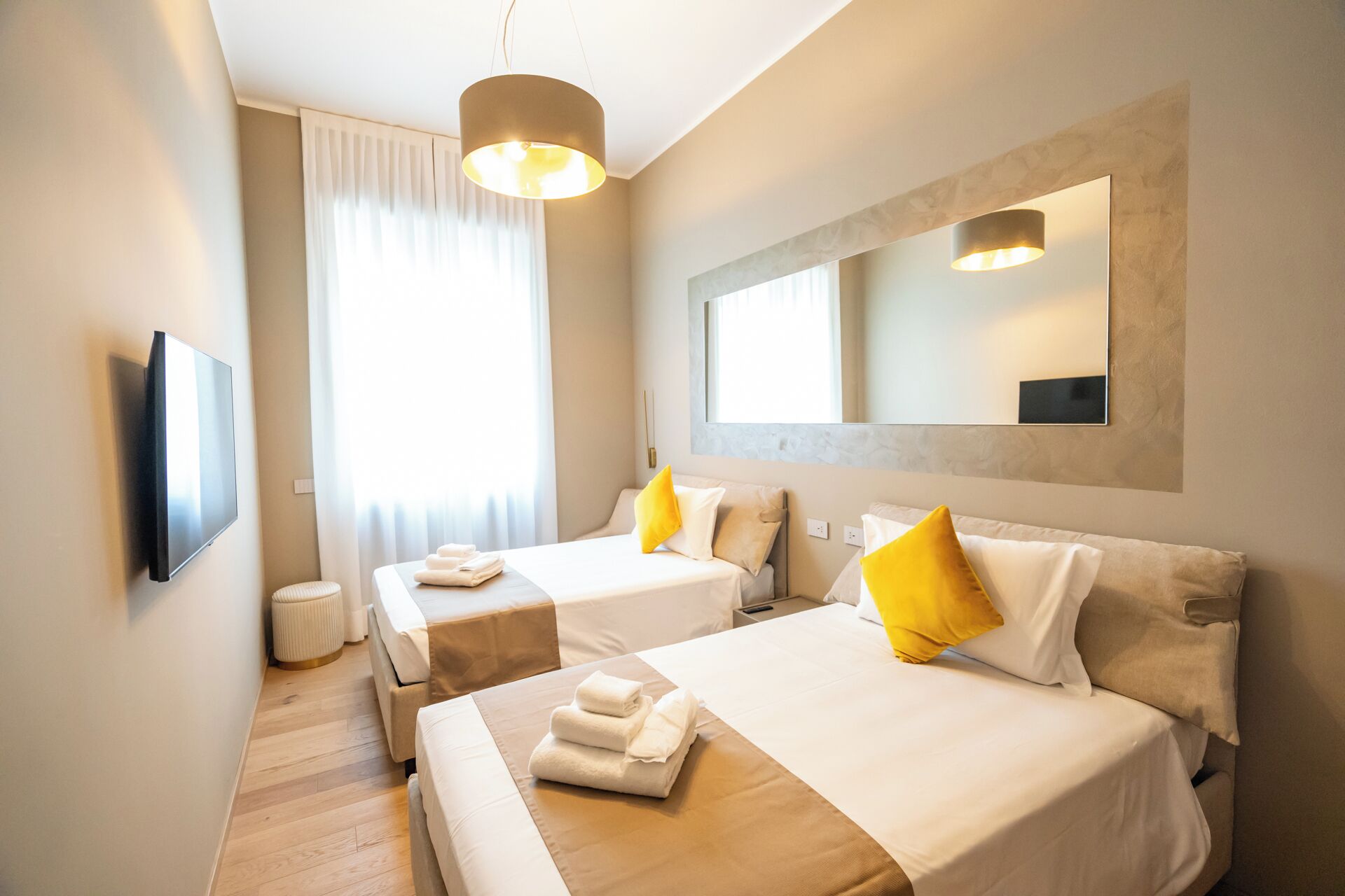 Une chambre d’hôtel avec deux lits, un téléviseur fixé au mur, un grand miroir et une fenêtre avec des rideaux.