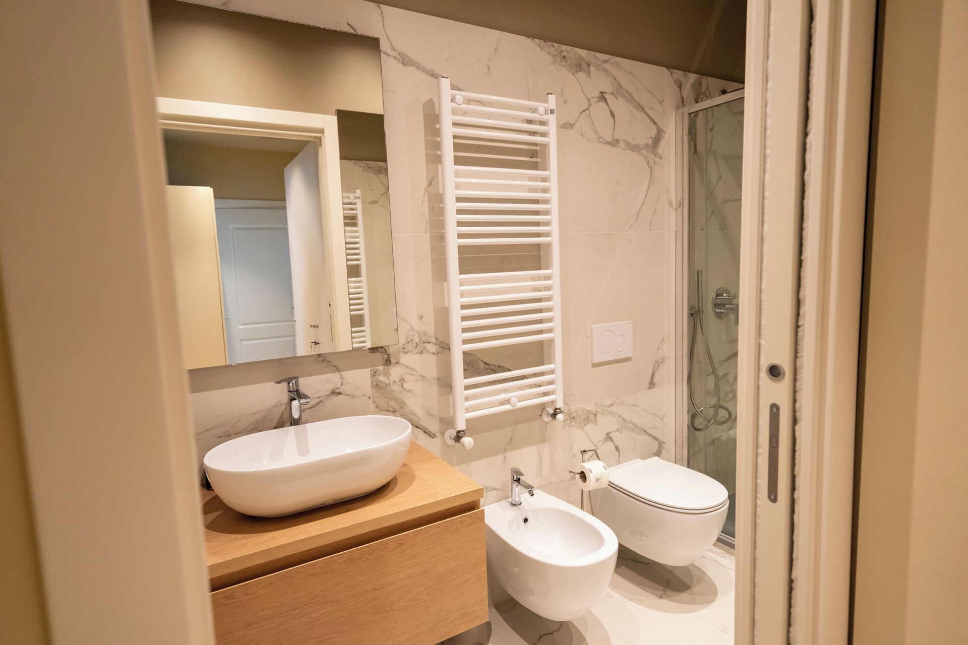 A modern bathroom with a white sink, bidet, and toilet, marble walls, and a glass shower enclosure.