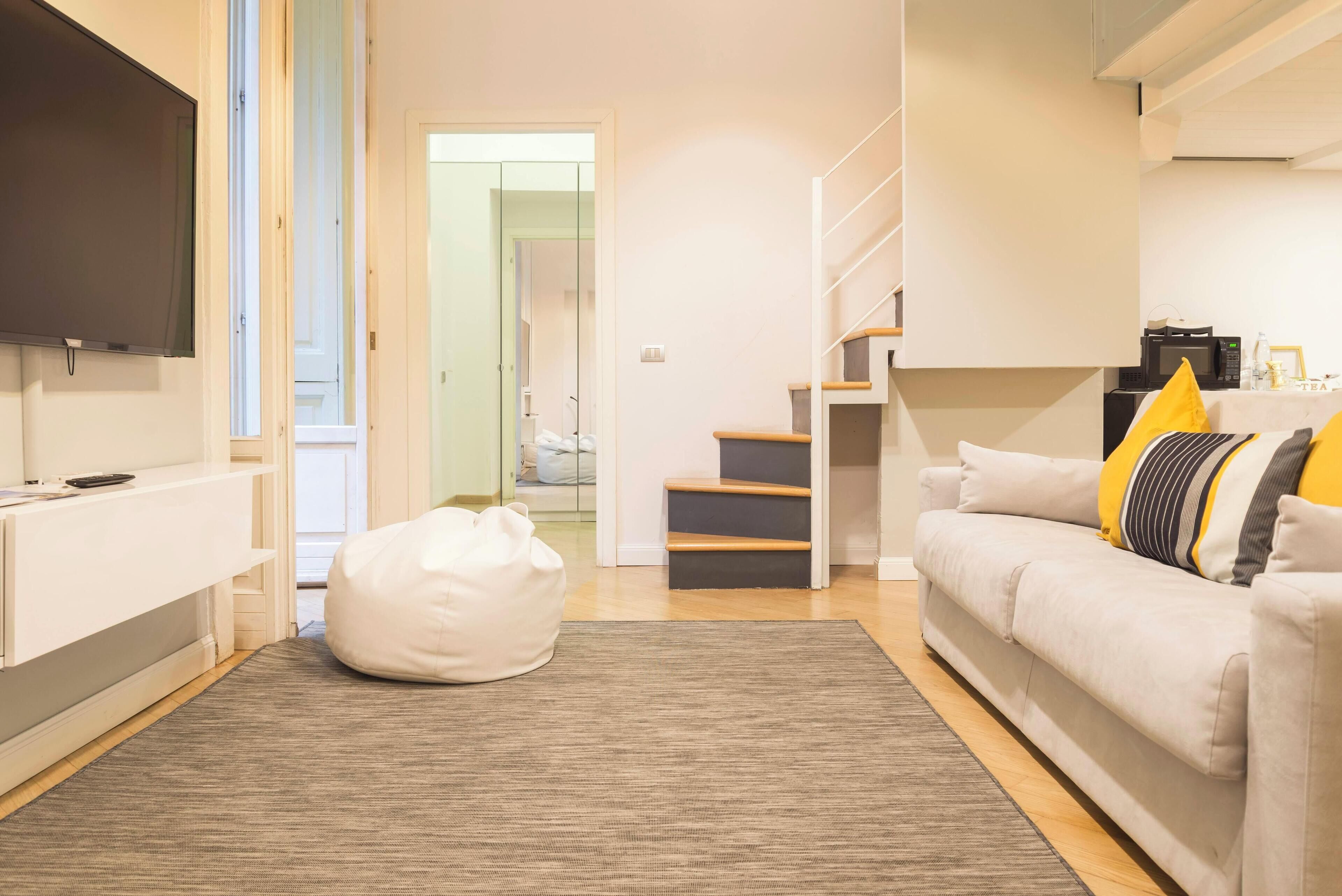Un salon moderne avec un téléviseur à écran plat, un fauteuil en forme de poire blanc, un tapis gris, un canapé blanc agrémenté de coussins jaunes et gris, et un escalier menant à un niveau supérieur.