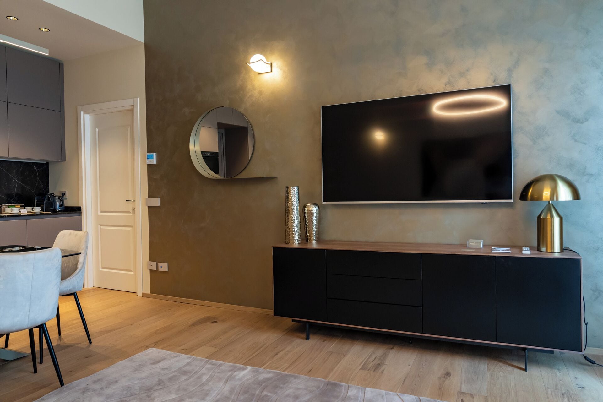 Ein modernes Wohnzimmer mit einem an der Wand befestigten Flachbildfernseher, einem runden Spiegel, einem schwarzen Konsolentisch mit Dekoration und einem hellen Sofa.