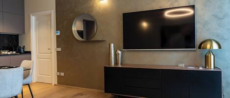 Un salon moderne avec un téléviseur à écran plat fixé au mur, un miroir rond, une console noire agrémentée d’objets décoratifs et un canapé de couleur claire.