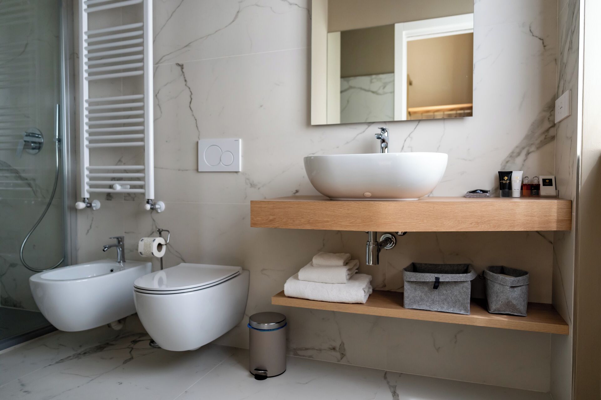 A modern bathroom with a marble floor, a white sink with a large basin, a bidet, a toilet, a mirror, and storage shelves.