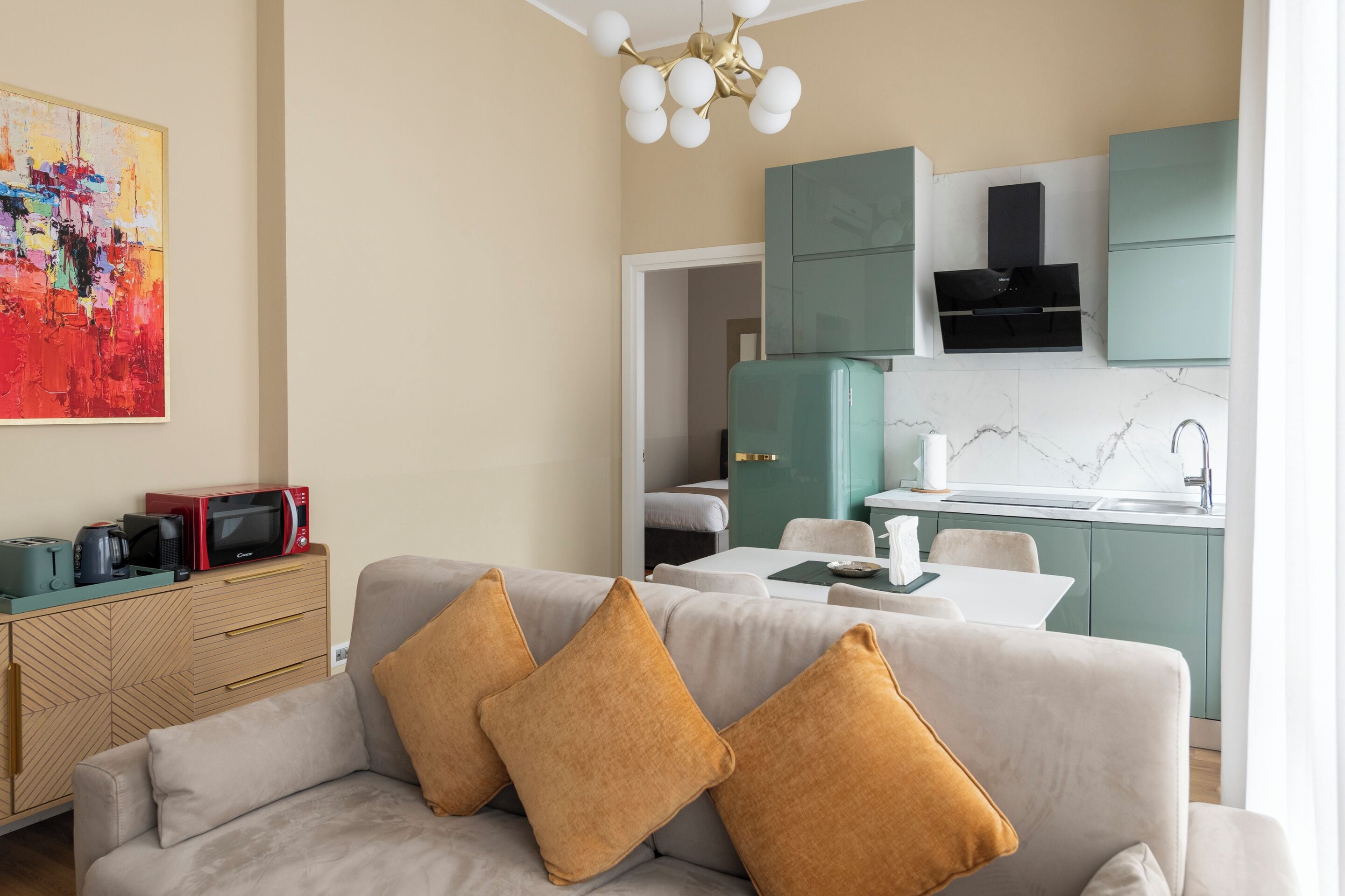 A modern living room with a sofa, a dining table, and a kitchen area with teal cabinets and a black range hood.