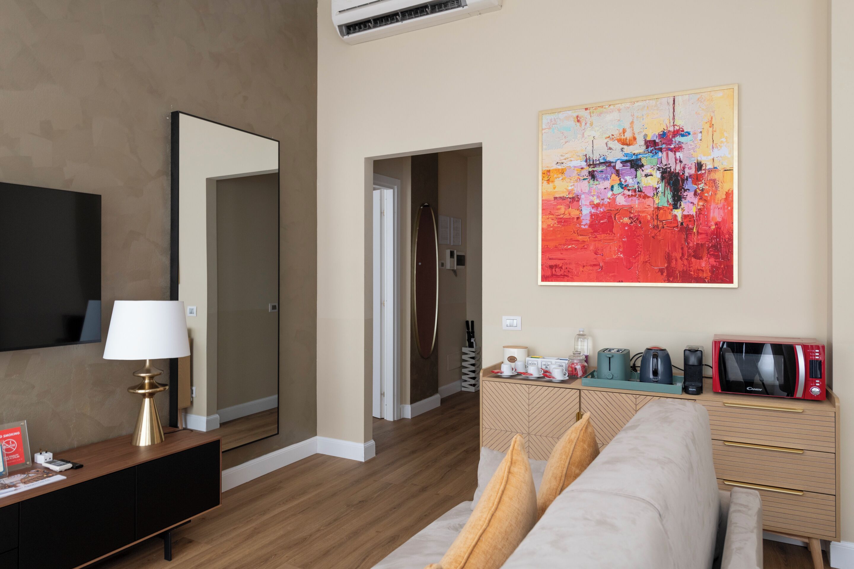 Habitación de hotel moderna con suelo de madera, un espejo grande, un televisor de pantalla plana, un sofá color beige, una mesita de centro y un cuadro abstracto y colorido en la pared.