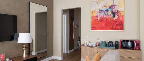 Habitación de hotel moderna con suelo de madera, un espejo grande, un televisor de pantalla plana, un sofá color beige, una mesita de centro y un cuadro abstracto y colorido en la pared.