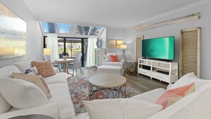 A spacious living room with a large TV, white furniture, and a view through a glass door to an outdoor area.