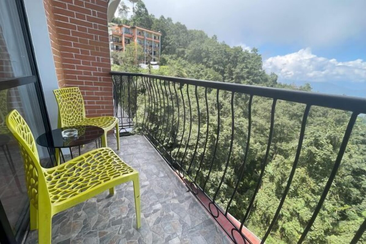 A balcony with yellow chairs and a table, overlooking a forested area and a building in the background.