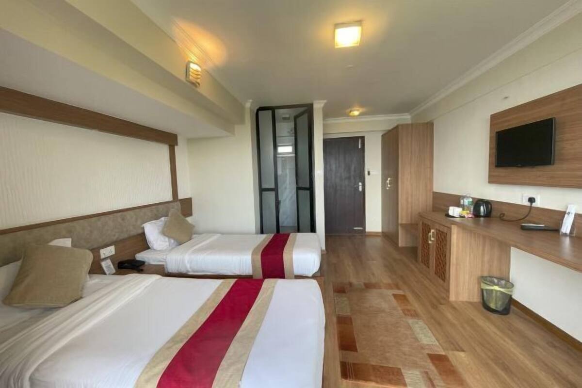A hotel room with two beds, a wooden floor, a TV mounted on the wall, and a small kitchenette area.