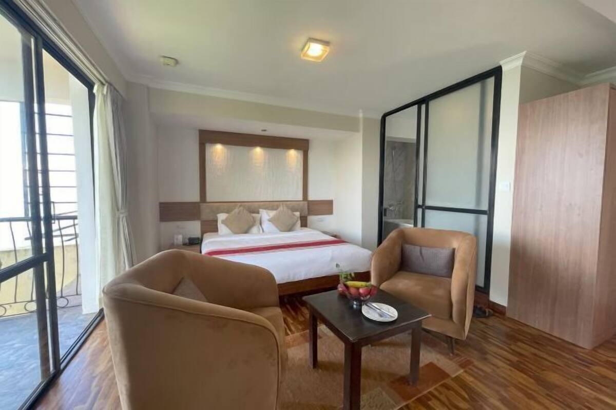 A hotel room with a bed, two armchairs, a small table with a fruit bowl, and a view of the outside.