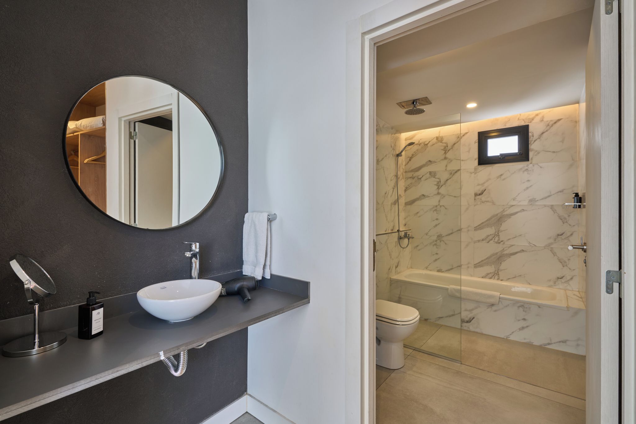 A modern bathroom with a round mirror, a white sink, a handheld showerhead, and a bathtub with marble walls.
