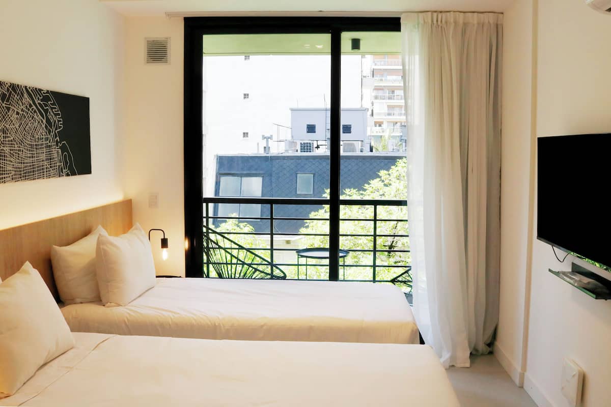 A hotel room with a large bed, a flat-screen TV, a window with a view of buildings, and a balcony with chairs.