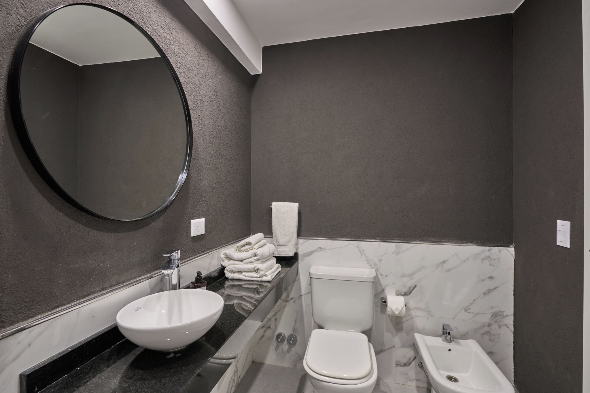 A modern bathroom with a round mirror, a sink, a toilet, and a bidet.