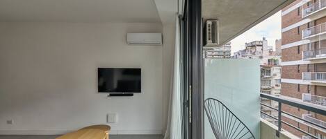 A balcony with a view of a brick building, a flat-screen TV mounted on the wall, a wooden table, a metal chair, and a sofa.