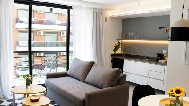 A modern living room with a sofa, a small table, and a chair. There is a large window with a view of a building outside. - Alma Rentals French Palermo (Buenos Aires)