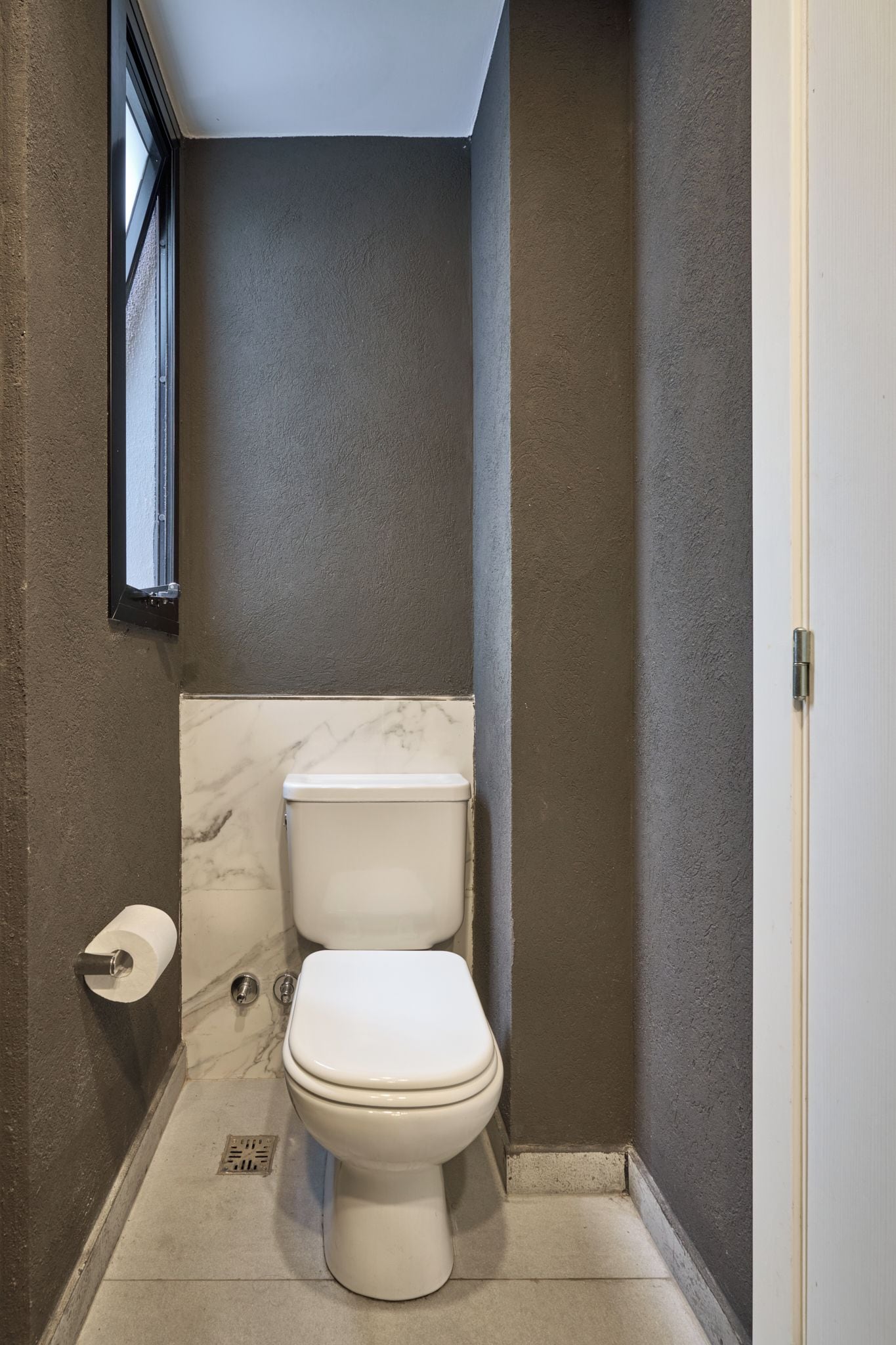 A small, compact bathroom with a toilet, a marble-tiled wall, and a window.