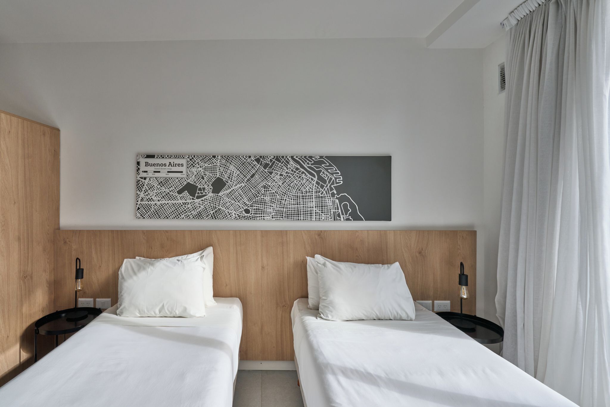 Een hotelkamer met twee bedden, een houten hoofdbord, een wereldkaart en een raam met gordijnen.