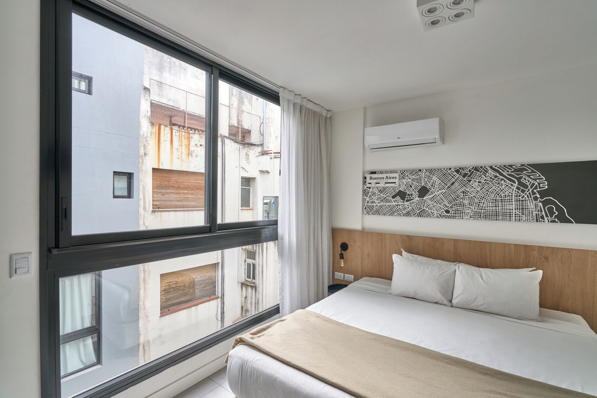A hotel room with a large window, a bed with white bedding, a wall-mounted air conditioner, and a cityscape view.