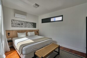 A modern hotel room with a large bed, a wooden headboard, a wall-mounted air conditioner, a map artwork, a window with a view, and a woven bench. - Alma Rentals French Palermo (Buenos Aires)