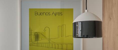 Una obra de arte con el cuadro de un paisaje urbano enmarcado, titulada 'Buenos Aires', en una pared, una mesa blanca con sillas y una lámpara colgante.