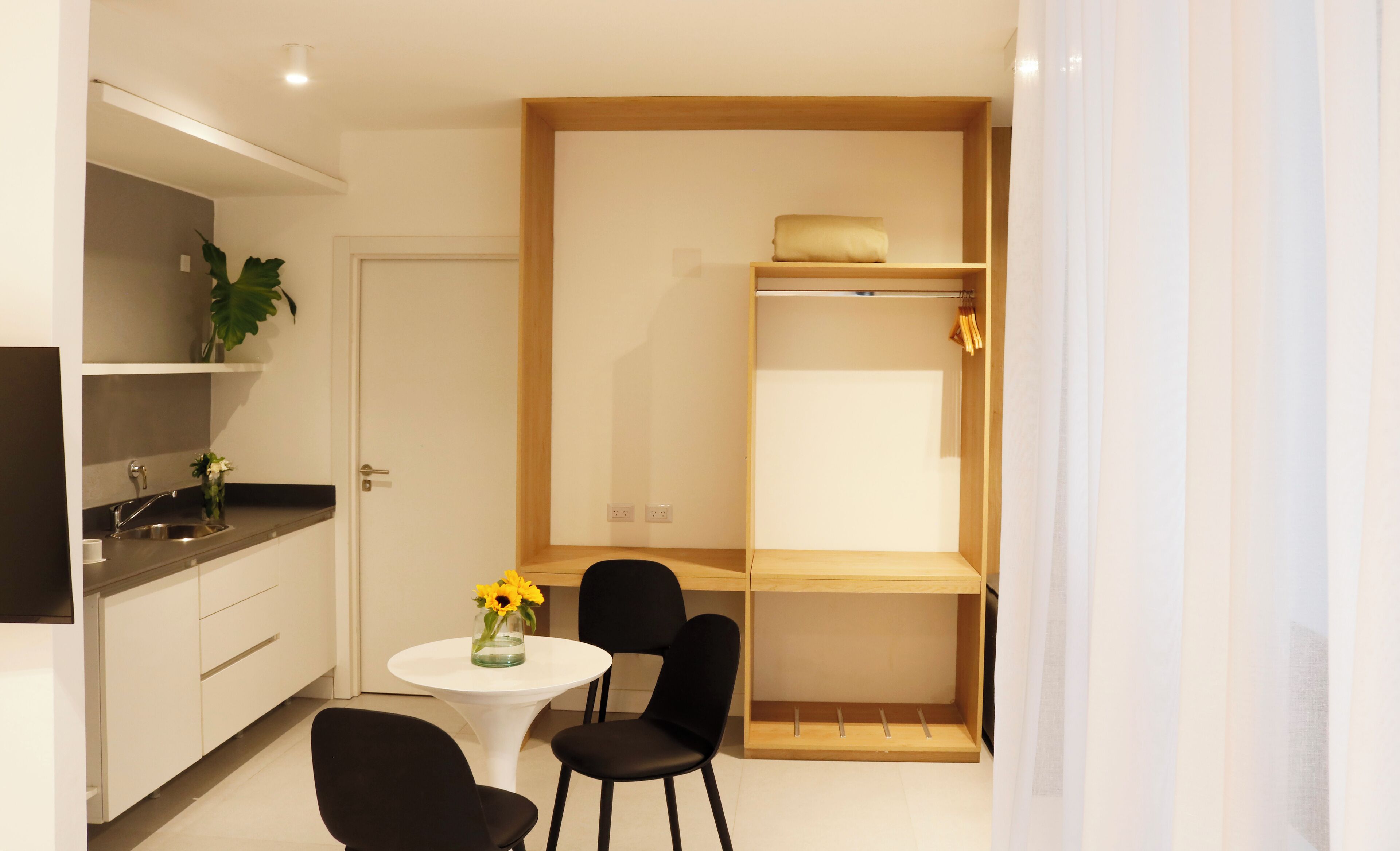Une cuisine compacte comprenant un évier, une petite table à manger et des chaises. Une étagère en bois est fixée au-dessus du plan de travail, et on y trouve une serviette et une plante.