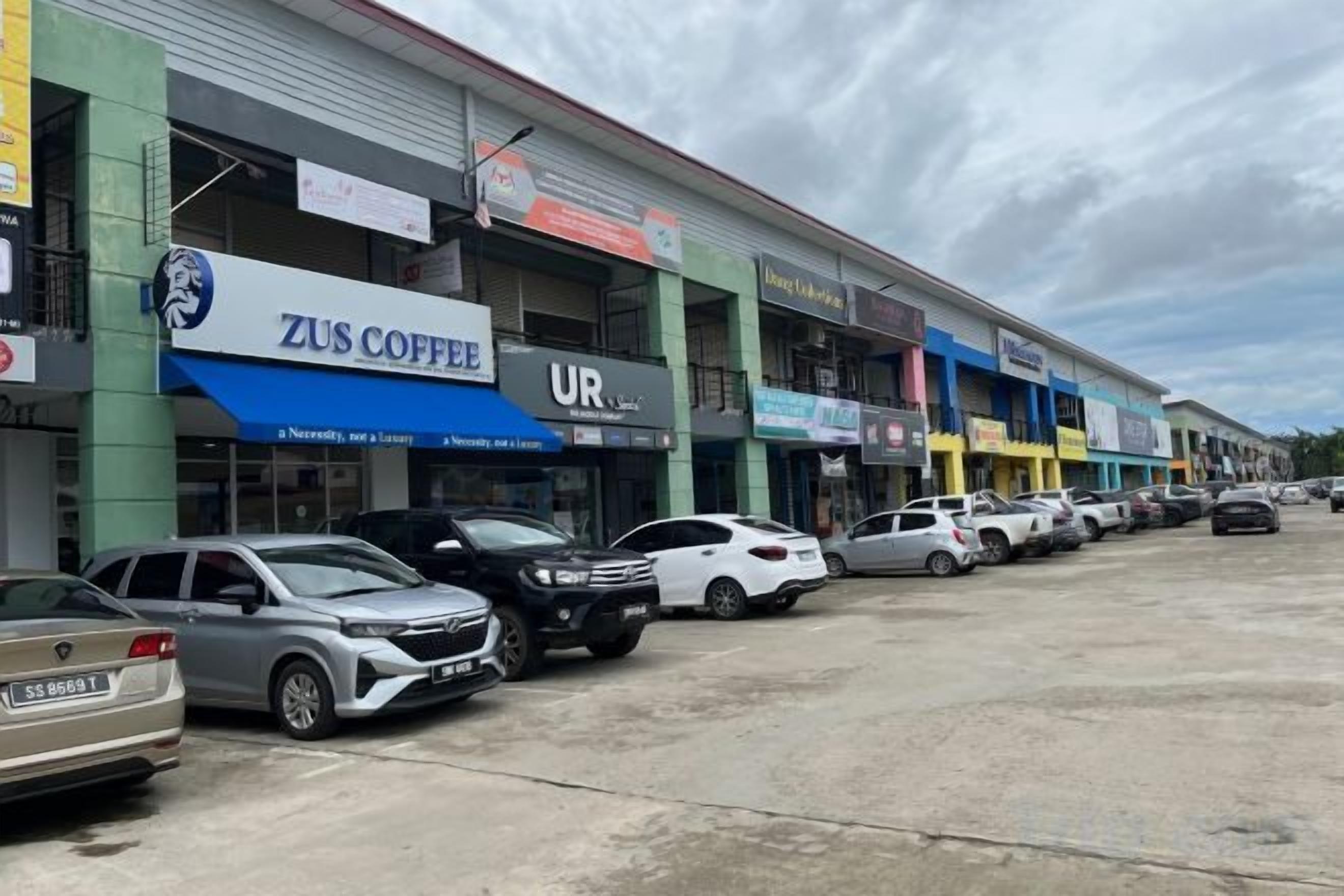 A commercial building with multiple storefronts, including a coffee shop named ZUS COFFEE, and a parking lot with several cars.