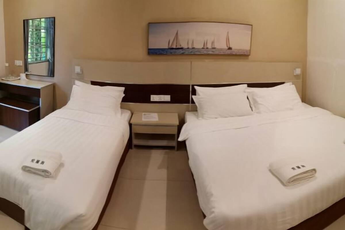 A hotel room with two single beds, a nightstand, a small table, a painting of sailboats, and a window with curtains.