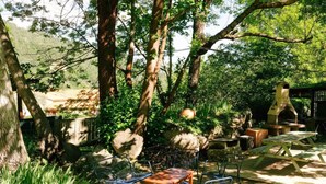 An outdoor patio with metal chairs and a wooden table, surrounded by trees and a stone fireplace.