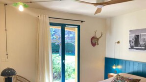 Un dormitorio con cama, una mesa de madera, una silla, un ventilador de techo y vistas al exterior a través de una puerta.