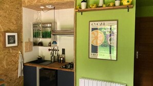 Ett litet, praktiskt kök med en inbyggd spis, en diskho och ett fönster med galler. På väggen hänger en inramad affisch, och ovanför den finns en hylla med köksredskap.
