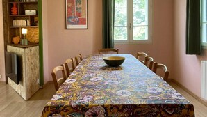 A dining area with a long table covered in a floral tablecloth, surrounded by chairs, a bookshelf with books and decorative items, and a window with curtains. - Studio (2 people) (SAHORRE)