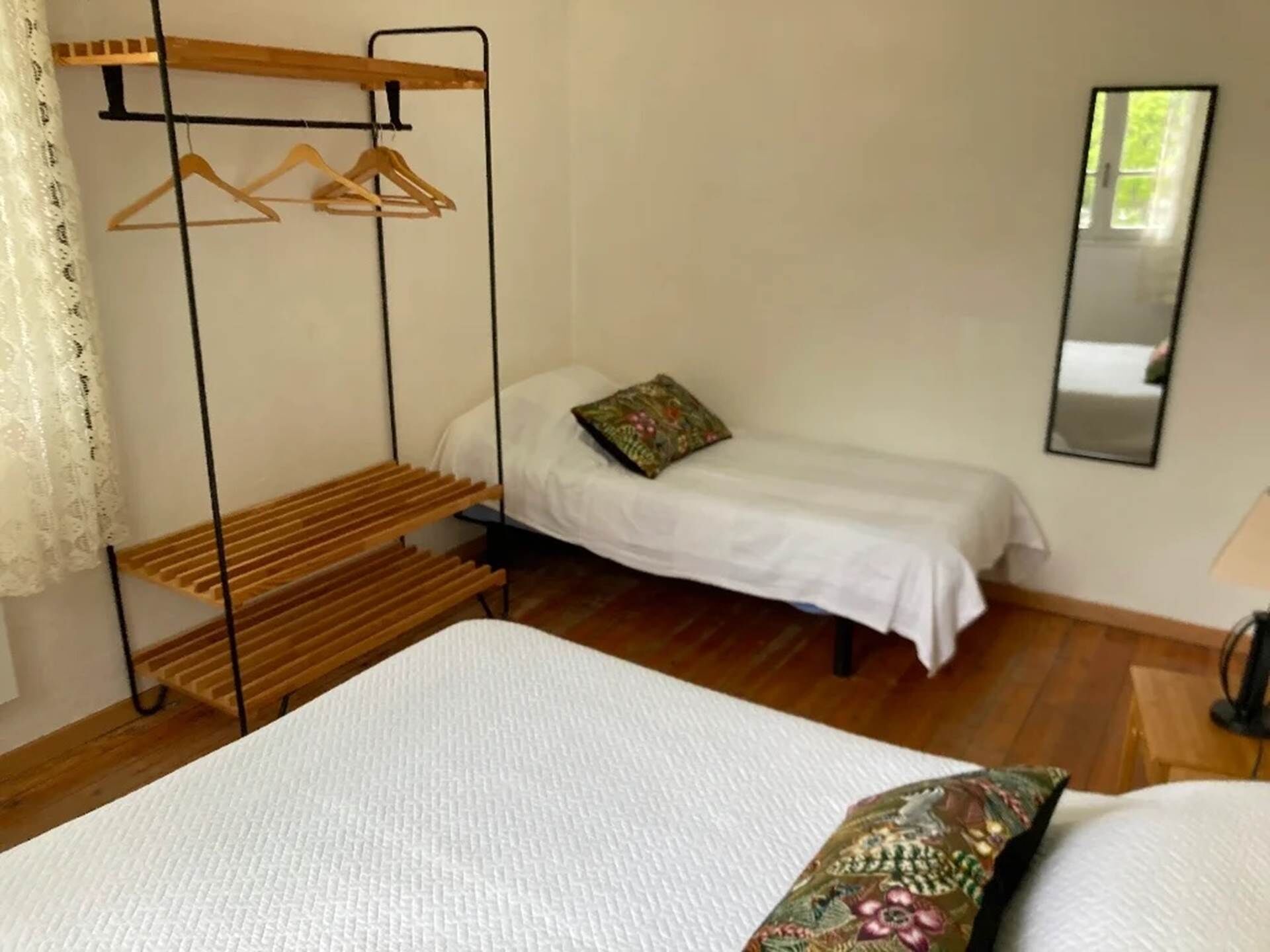 A bedroom with a single bed, a wooden drying rack, a small bedside table, and a mirror on the wall.