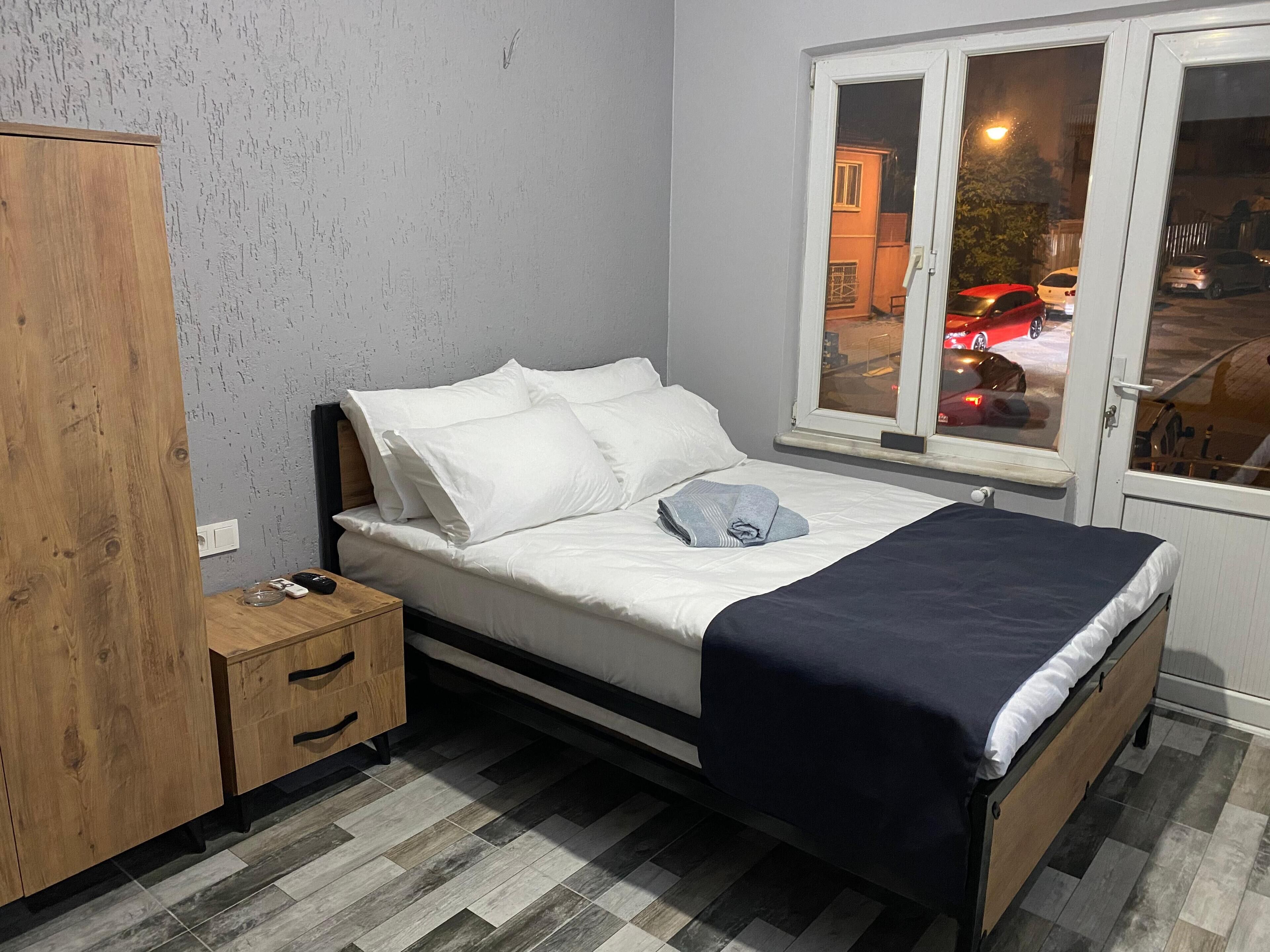 A bedroom with a bed, a wooden wardrobe, a nightstand, and a window with a view of the street.
