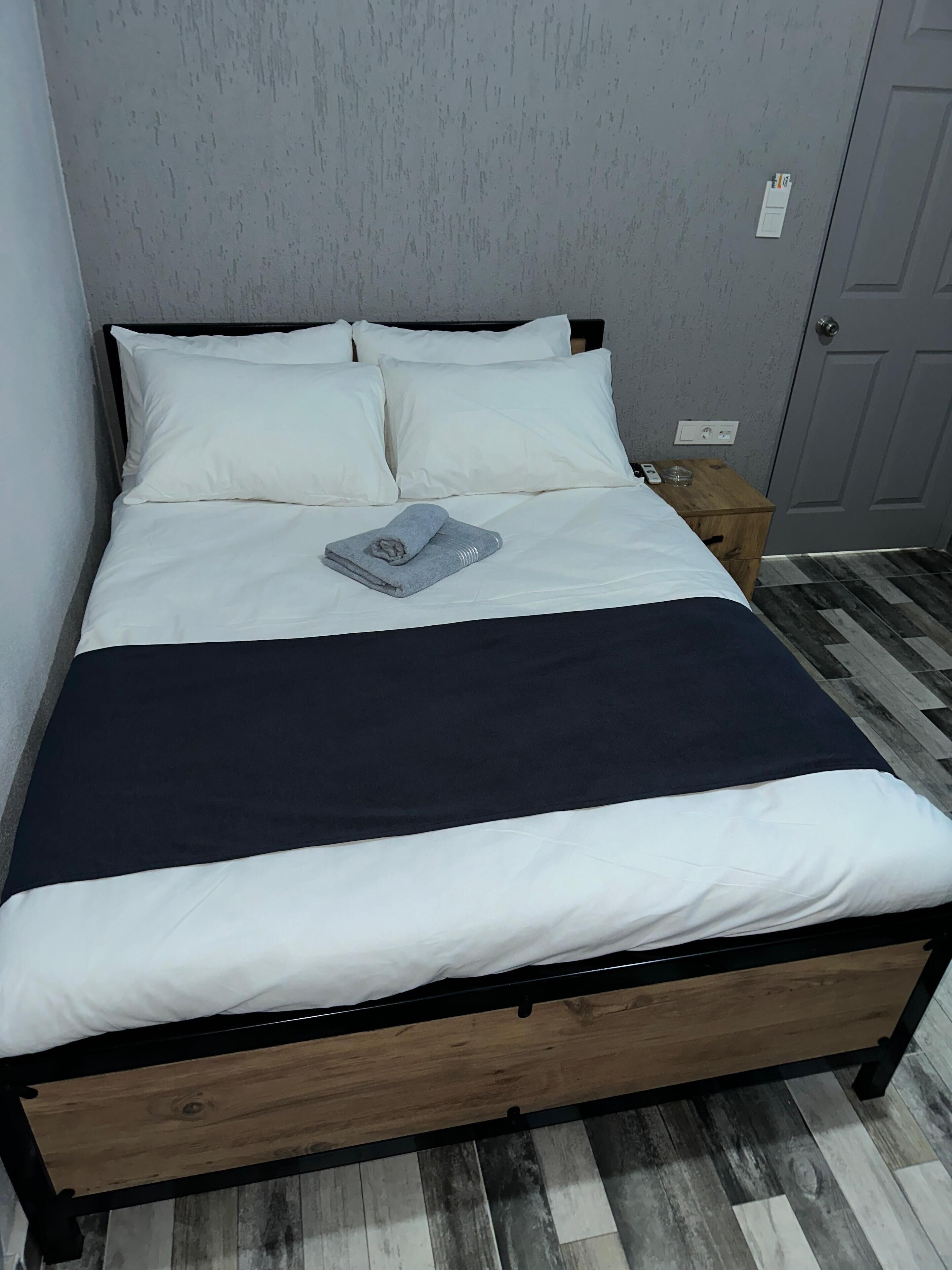 Un letto rifatto con lenzuola bianche e blu scuro, un asciugamano piegato e un comodino con un bicchiere e un telefono.