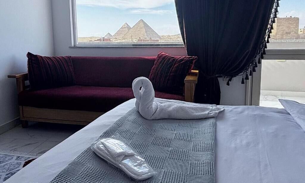 Quarto de hotel com uma cama, um sofá vermelho, uma janela com vista para pirâmides e uma cortina.