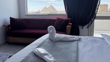 Quarto de hotel com uma cama, um sofá vermelho, uma janela com vista para pirâmides e uma cortina.