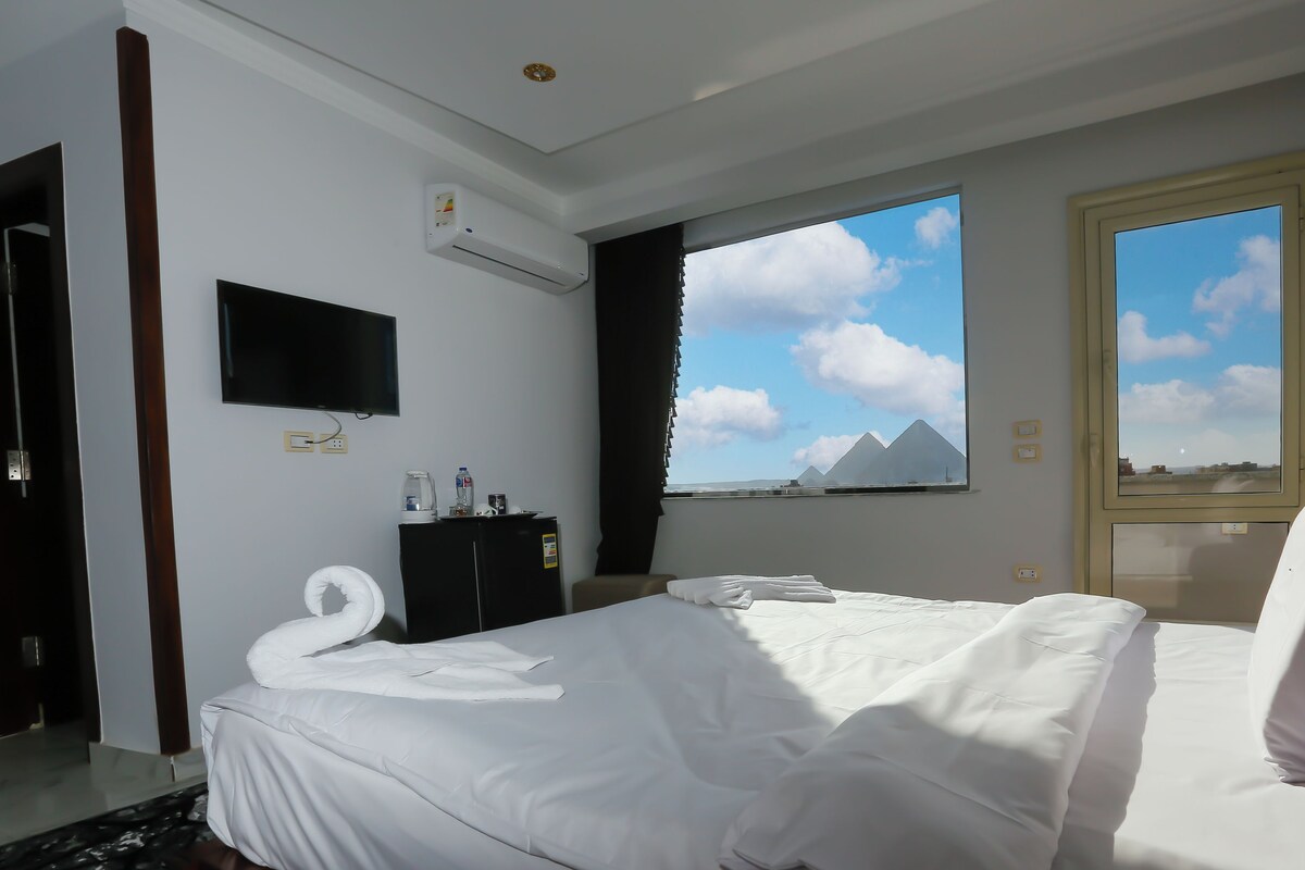 A hotel room with a bed, a flat-screen TV, a minibar, a window with a view of pyramids, and a door.