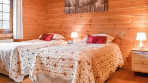 A wooden cabin bedroom with two beds, floral bedding, a painting on the wall, and a window with curtains.