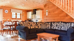 A cozy cabin interior with a wooden dining table, chairs, a sofa with patterned cushions, and a staircase.