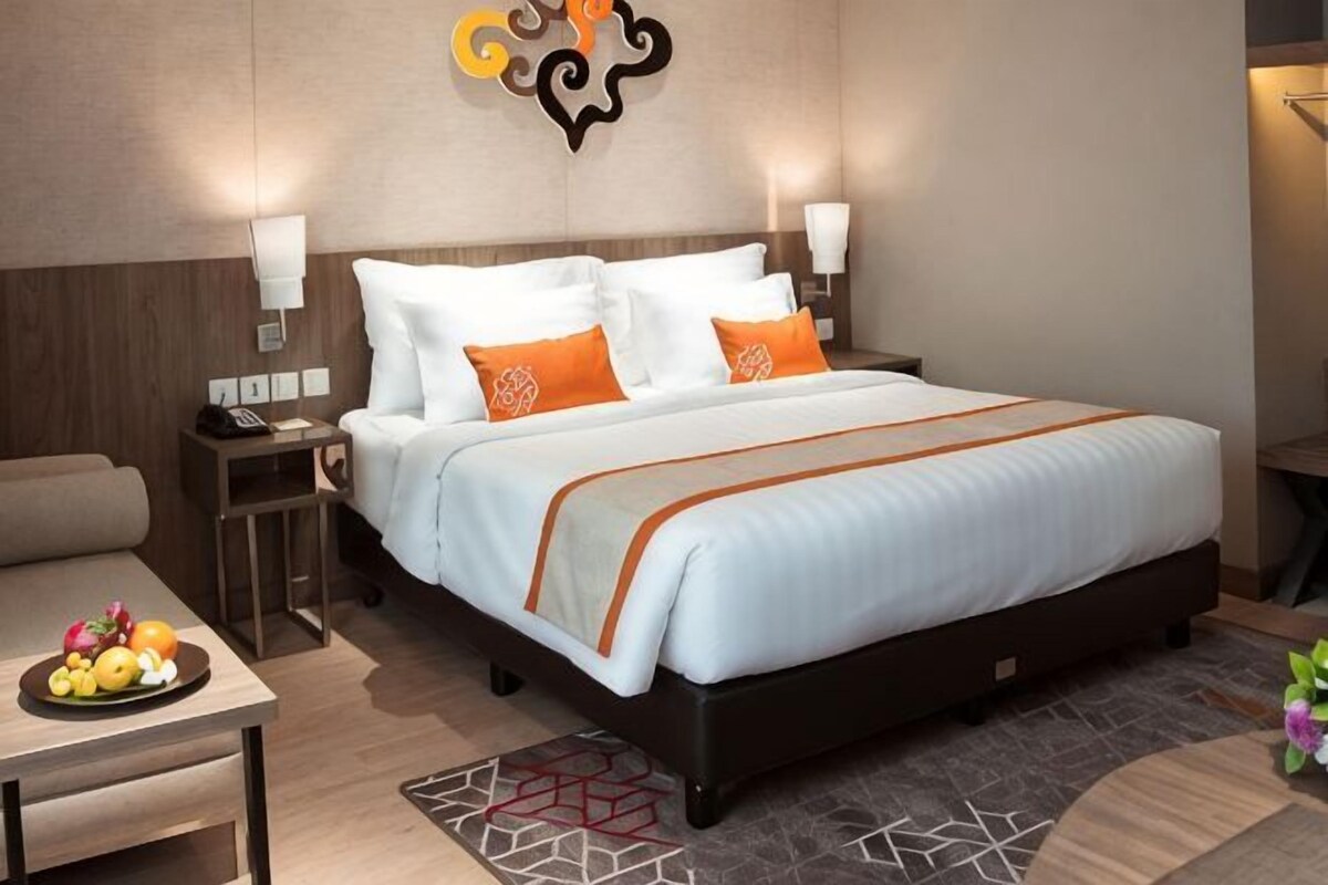A hotel room with a large bed, two bedside tables, a nightstand, a desk with a fruit bowl, and a decorative wall piece.