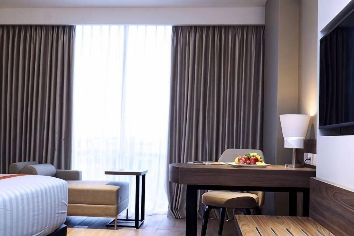 Quarto de hotel com cama, escrivaninha, cadeira, abajur e vista através de grandes janelas.