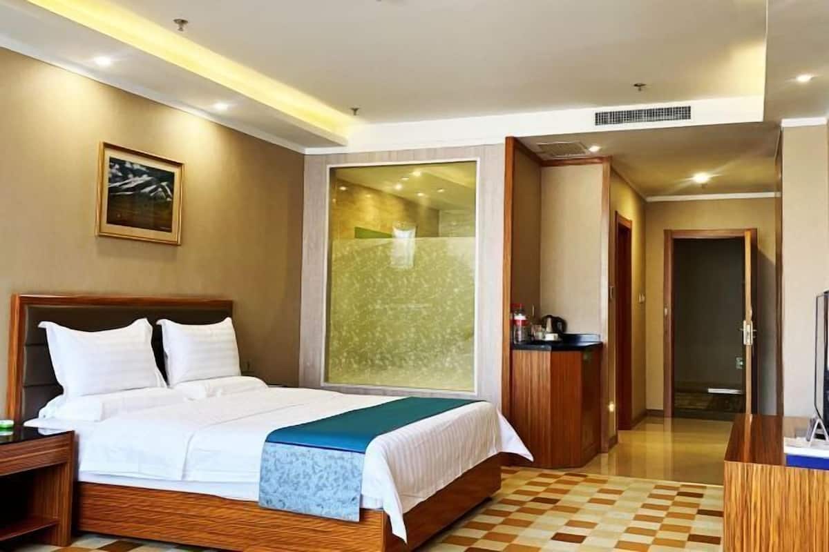 A hotel room with a bed, a nightstand, a desk, a television, and a bathroom visible through a glass partition.
