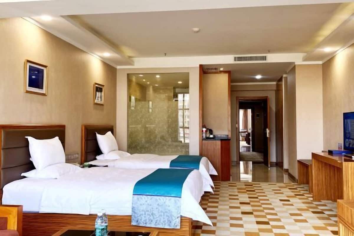 A hotel room with two beds, a desk, a chair, a television, and a bathroom visible through a glass door.