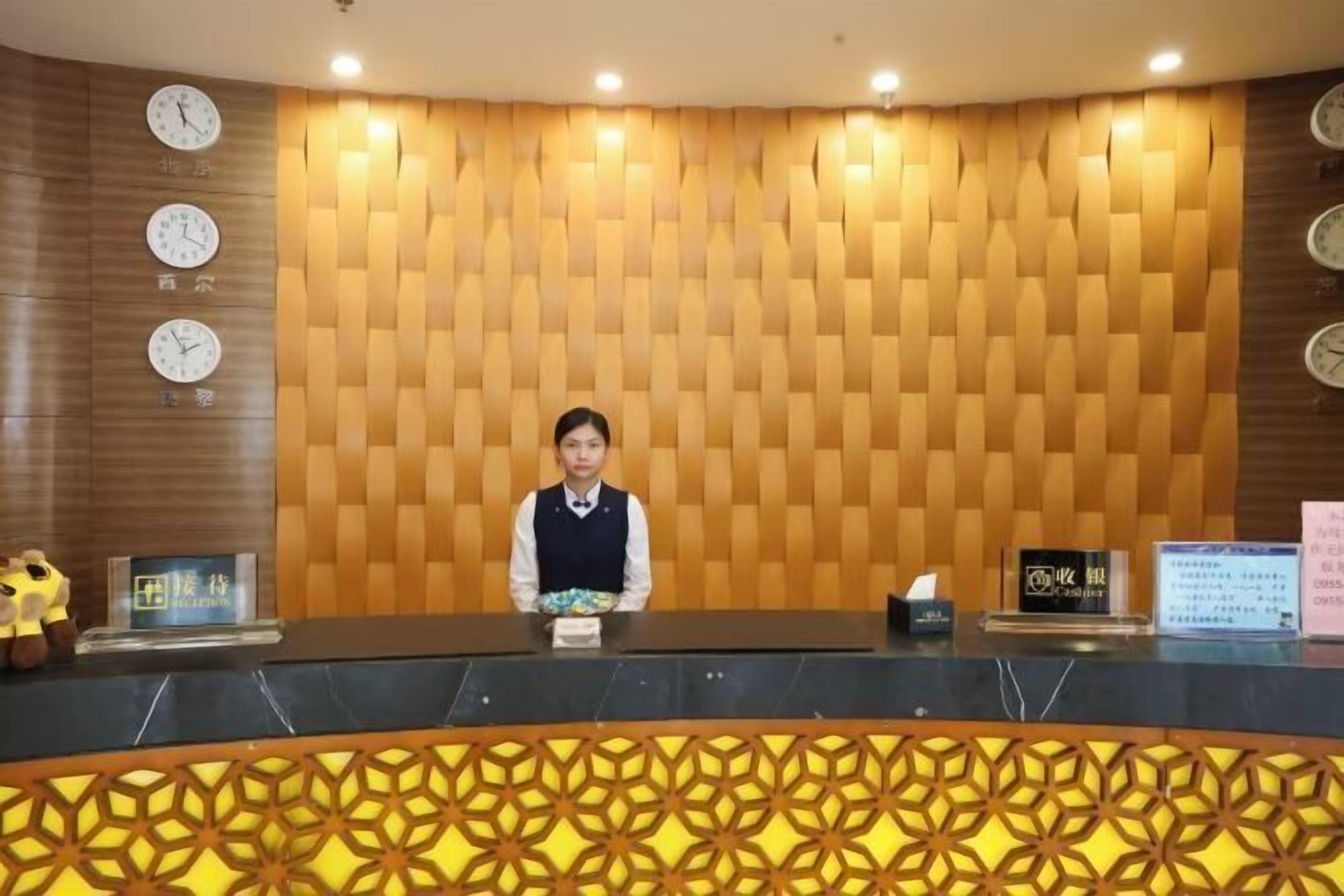A reception desk with a patterned wall, clocks, and a person seated behind it.