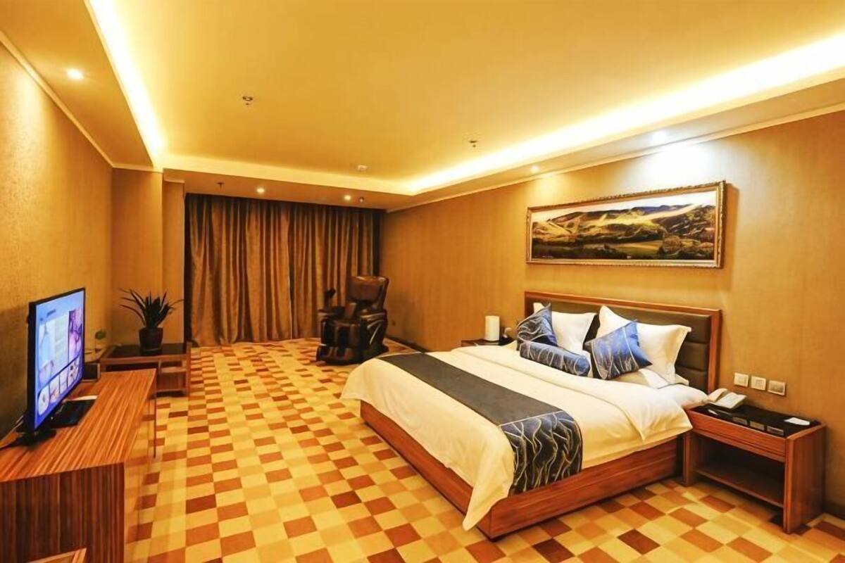 A hotel room with a checkered floor, a bed with multiple pillows, a desk with a television, and a painting on the wall.