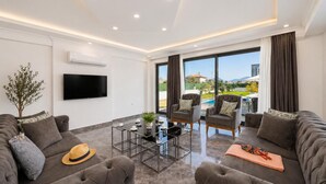 Una sala de estar moderna con un televisor de pantalla plana, sofás grises, una mesa de centro y vistas a una piscina y montañas.
