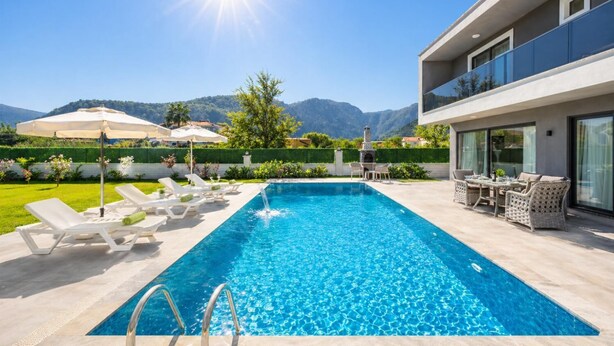 Piscina con sillas de descanso y una mesa, rodeada por un edificio moderno y vistas a montañas.