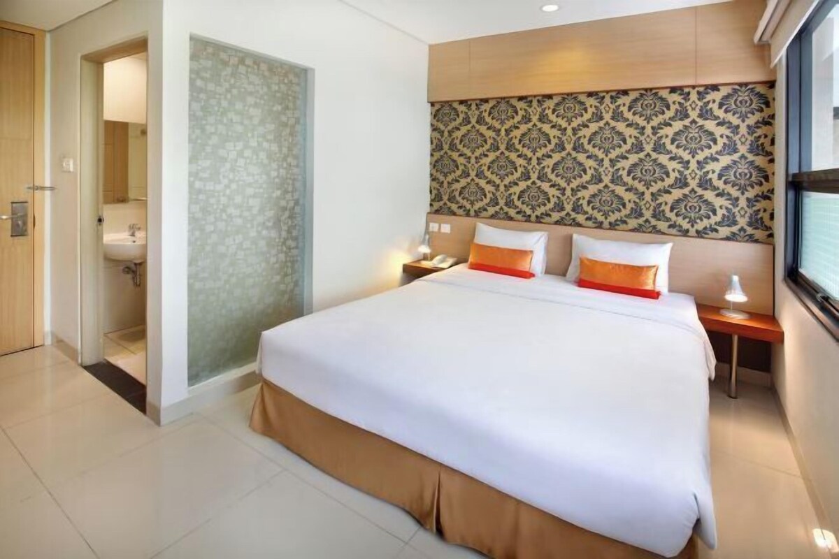 A hotel room with a large bed, a nightstand, a bedside table, a bathroom with a toilet and sink, and a patterned wall.
